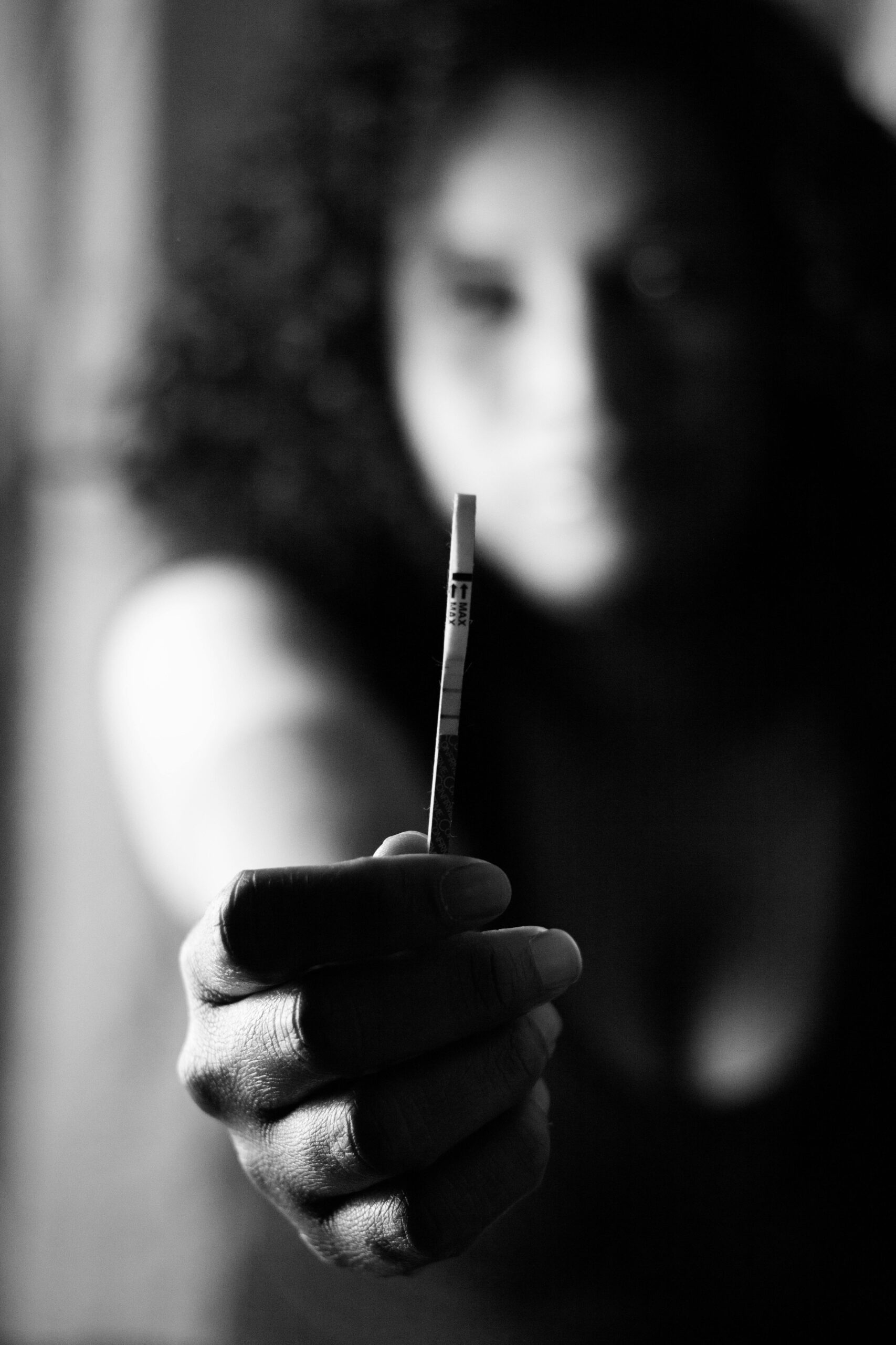 A woman's hand holding a pregnancy test strip in focus, monochrome.